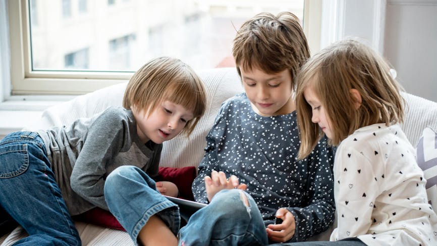 Trois enfants de plus de 3 ans jouent avec une tablette sur un canapé